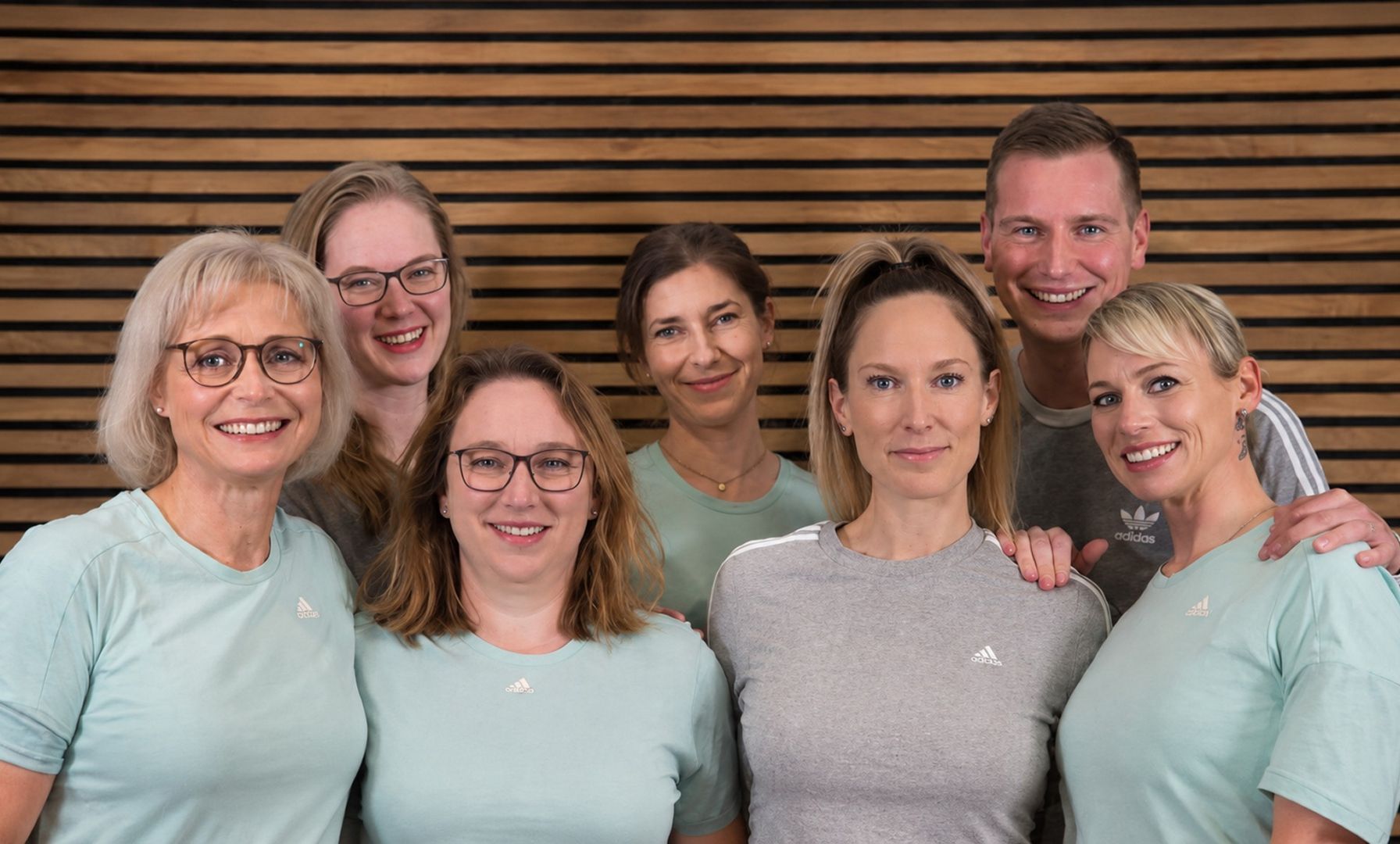 Sieben lächelnde Personen in Adidas-Sportkleidung vor einer Holzwand-Kulisse